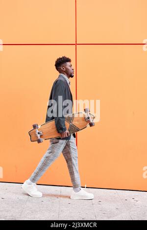 Junger Mann mit Blazer und Skateboard, der vor orangefarbenem Hintergrund die Straße entlang läuft. Stockfoto