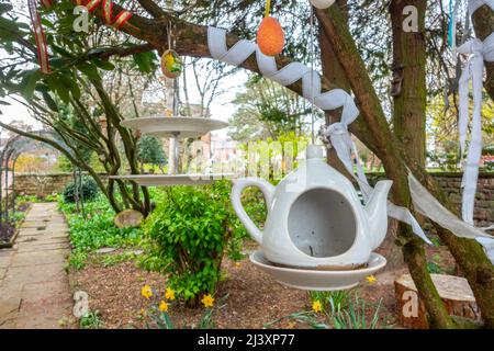 Eine Teekannen- und Kuchengarbe, die in einem öffentlichen Garten in Penrith, Cumbria, Großbritannien, an Baumzweigen hängen, ähneln der Teeparty von Mad Hatter Stockfoto