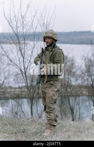 Ukrainischer Soldat steht mit kalaschnikow-Sturmgewehr in den Händen vor dem Hintergrund von Himmel und Fluss. Konzept der russischen Militärinvasion in Ukra Stockfoto