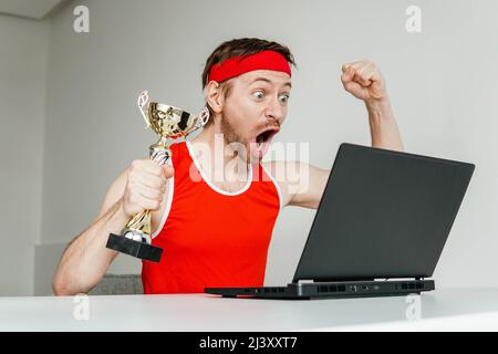 Erfolgreicher Sportler in roter Sportbekleidung feiert den Sieg mit der goldenen Pokal-Trophäe und beobachtet sein Ergebnis auf dem Laptop. Stockfoto