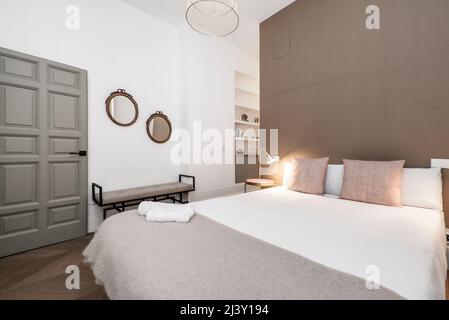 Schlafzimmer mit Ankleidezimmer und eigenem Bad hinter der Wand des Kingsize-Bettes mit rötlichen Kissen und passendem Hocker Stockfoto