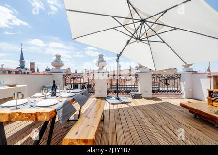 Terrasse mit großem weißen Stoffschirm mit Holzböden, Esstisch aus Massivholz mit Bänken und passendem Beistelltisch auf einer städtischen Terrasse mit einem Vin Stockfoto