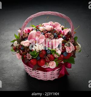 Rosafarbener Weidenkorb mit Erdbeeren, Heidelbeeren, Kirschen und rosa Blüten auf schwarzem Hintergrund Stockfoto