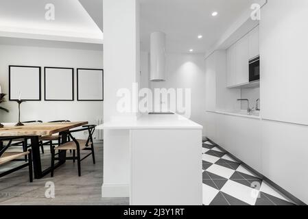 Designer-Küche mit weichen weißen Linien, integrierten Geräten und weiß lackierten Möbeln. Esstisch aus Maiza-Holz und kombinierte Steinzeugböden Stockfoto