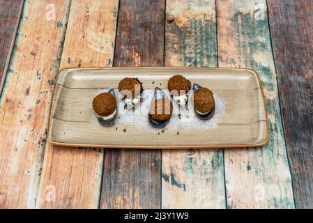 Köstliche Garnelenkroketten serviert Marinara-Stil auf Muschelschalen, grobem Salz, Schnittlauch, Mayonnaise und einem beigen Teller auf einem Holztisch Stockfoto