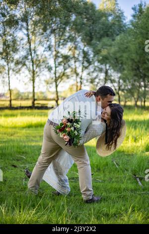 Glückliches Hochzeitspaar, der Bräutigam bewundert die glückliche Braut, hält die Braut in den Armen und wirbelt sie, an einem sonnigen Sommertag im Park, fröhlich zur Hochzeit Stockfoto