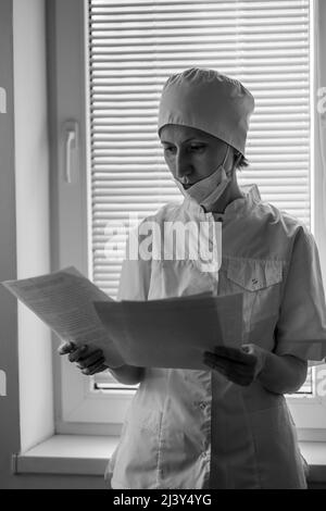 Die Ärztin steht in der Nähe des Krankenhausfensters und schaut durch die Dokumente. Schwarzweiß-Foto. Stockfoto