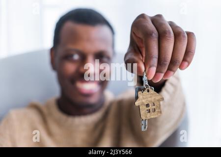 Afroamerikanischer Mann prahlt mit dem Kauf einer neuen Wohnung. Fokus ändert sich von Schlüssel mit Holzhaus Trinket zu glücklichen Hausbesitzer close view Stockfoto