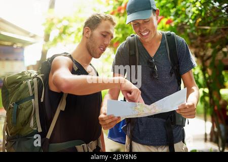 Den Weg zu einer neuen Distination finden. Aufnahme von zwei lächelnden jungen Freunden, die in Rucksäcken unterwegs sind und sich während ihres Aufenthalts in Thailand über eine Karte unterhalten. Stockfoto