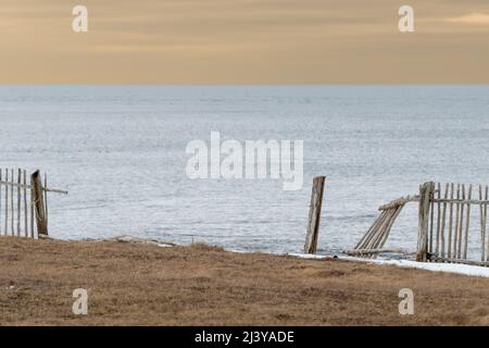 Ein gebrochener Zaun am Rand einer Klippe mit Blick auf das Meer. Der Holzzaun besteht aus Holzstäben. Das Meer ist ruhig und blassblau und himmelgelb. Stockfoto