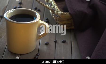 Eine hellbraune Tasse gefüllt mit schwarzem Kaffee auf einer Holzdiele-Tischplatte mit verstreuten Kaffeebohnen neben einem Korbtablett, das mit dunkelbraunem Leinen bedeckt ist. Stockfoto