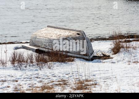 Ein weißes Fischerboot aus Holz mit abblätternder Farbe, das neben dem Meer und der felsigen Küste mit Schnee auf dem Boden befahren wurde. Das offene Boot ist von unten nach oben. Stockfoto