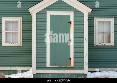 Die Außenfassade eines alten Holzgebäudes. Es gibt eine grüne Einzeltür mit rostigen Scharnieren, weißen Zierleisten und Holzbrettern auf dem Haus. Stockfoto