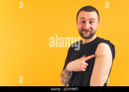 Impfkonzept. Glücklicher Gangster-ähnlicher bärtiger Kaukasier mit Tattoos, die nach der Impfung mit dem Finger auf seinen Arm zeigen und mit Pflaster bedeckt sind. Hochwertige Fotos Stockfoto