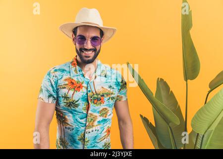 Gut aussehender hispanischer Mann in legerer Sommerkleidung, der neben grünen Blättern steht und mit dem Sommerurlaubskonzept Medium Shot Studio Shot mit orangefarbenem Hintergrund genießt. Hochwertige Fotos Stockfoto
