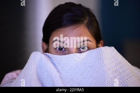 Was wird als nächstes passieren? Aufnahme einer jungen Frau, die ein Gesicht mit einer Decke bedeckt, während sie zu Hause fernsehen. Stockfoto