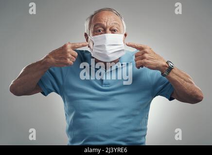 So trägst du eine Gesichtsmaske. Aufnahme eines älteren Mannes, der eine schützende Gesichtsmaske in einem Studio vor grauem Hintergrund trägt. Stockfoto