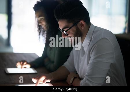 Konzentriert sich darauf, die Arbeit zu erledigen, egal was passiert. Eine kurze Aufnahme von zwei Kollegen, die spät in einem Büro an ihren digitalen Tablets arbeiten. Stockfoto