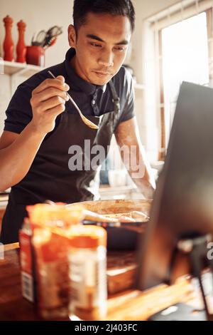 An einem Kochkurs von zu Hause aus teilnehmen. Aufnahme eines jungen Mannes, der ein Notebook benutzt, während er zu Hause eine Mahlzeit zubereitet. Stockfoto