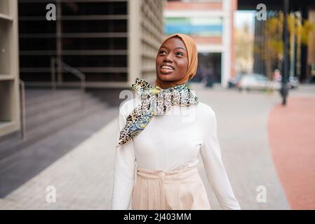 Positive, gut gekleidete afroamerikanische Frau im islamischen Kopftuch, die im modernen Stadtteil steht und mit einem breiten Lächeln wegschaut Stockfoto