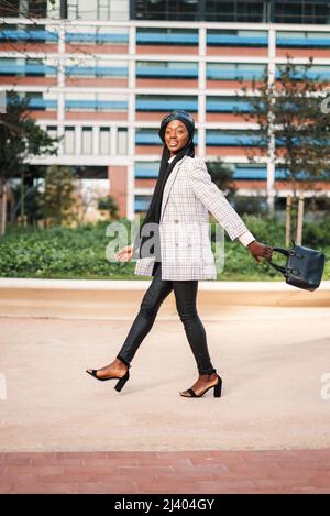 Seitenansicht Ganzkörper von sorglosen afroamerikanischen Frauen in trendigen formellen Outfit und muslimischen Kopftuch genießen Spaziergang im Stadtpark und lächeln glücklich Stockfoto