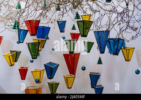 Bunte Glasoobjekte, die am Baumzweig hängen, mit weihnachtsornamenten und Lichtern an der weißen Wand Stockfoto