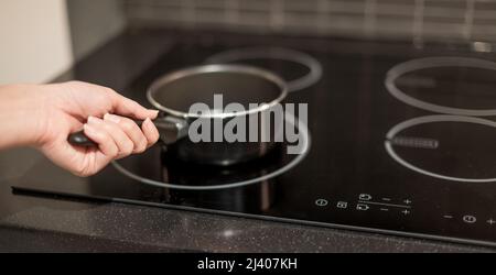 Weibliche Hand hält einen Topf, der in der Küche auf die elektrische Pfanne gelegt wird. Stockfoto