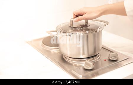 Weibliche Hand hält einen Topf, der in der Küche auf die elektrische Pfanne gelegt wird. Stockfoto