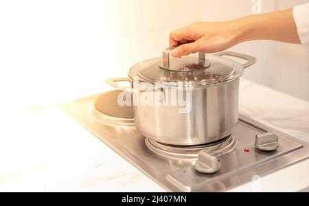 Weibliche Hand hält einen Topf, der in der Küche auf die elektrische Pfanne gelegt wird. Stockfoto