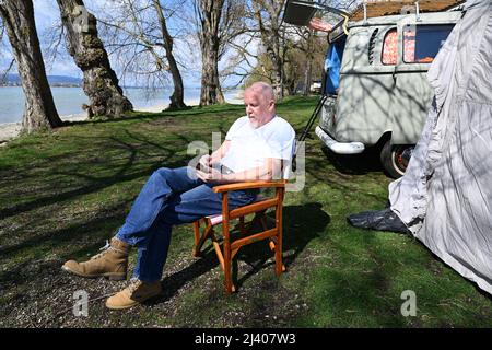 Allensbach Am Bodensee, Deutschland. 10. April 2022. Der Freiburger Camper Uwe sitzt in einem Campingstuhl direkt am Ufer des Bodensees neben seinem VW-Bulli (T2), nachdem er seine Markise in rauer Form aufgestellt hat. Wer Ostern im Land zelten will, muss sich beeilen: Der Landesverband der Campingunternehmer in Baden-Württemberg erwartet bei schönem Wetter einen ersten Ansturm auf die Ferien. Quelle: Felix Kästle/dpa/Alamy Live News Stockfoto