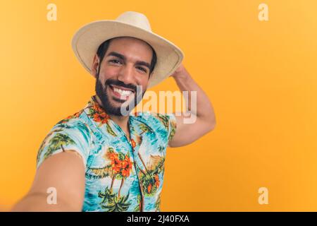 Weitwinkelansicht eines jungen lateinischen Mannes in Sommerkleidung, der seinen Arm ausstreckte und ein Selfie machte, während er seinen Hut hielt. Hochwertige Fotos Stockfoto