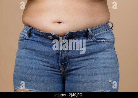 Kaukasische Dame zeigt großen Bauch mit großen übergewichteten Seiten in blauen Jeans. Viszerales Fett. Körper positiv und sich selbst akzeptieren. Plötzliche Gewichtszunahme Stockfoto