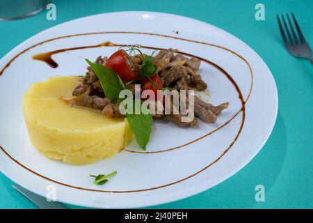 Fleischbällchen mit Kartoffelpüree auf einem weißen Teller. Fleisch in saurer Rahmsoße. Tomaten und Basilikum. Essen im Restaurant. Mittagspause. Medaillons. Stockfoto