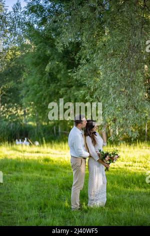 Braut und Bräutigam bei einem Spaziergang im Freien am Hochzeitstag. Brautpaar, glückliche junge Frau und Mann umarmen sich im grünen Park. Liebevolles Hochzeitspaar im Freien Stockfoto