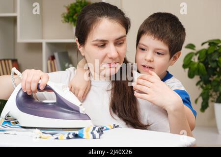 Ein kleines Kind von 4 Jahren hindert seine Mutter daran, Kleidung zu bügeln. Die Schwierigkeiten der Mutterschaft. Stockfoto