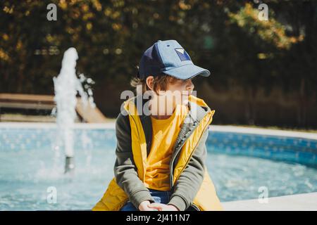 Schuljunge, der an einem sonnigen Frühlingstag auf einem Brunnen im öffentlichen Park sitzt und nachdenklich zur Seite schaut. Kind in gelber Weste, das im Park mit Wasser chillend ist Stockfoto