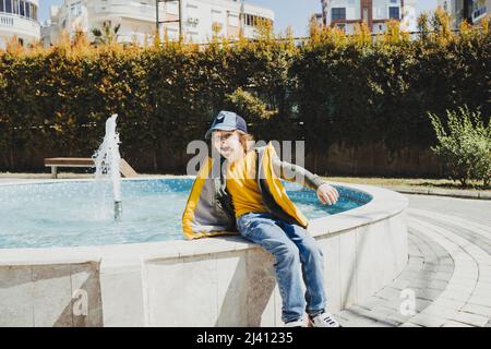 Schuljunge, der an einem sonnigen Frühlingstag auf einem Brunnen im öffentlichen Park sitzt und mit Wasser spielt. Kind in gelber Weste, das im Park mit Wasser bestreut chillt Stockfoto