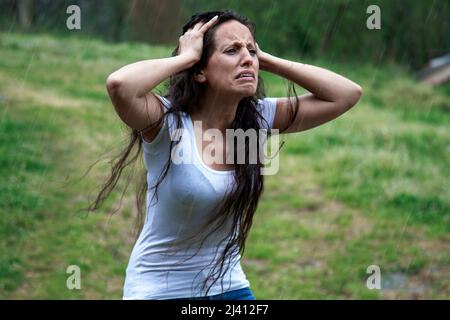 Verzweifelte Frau, die im Regen ihre Hände am Kopf hält. Weinend und bekümmert, schlechte Nachrichten. Trauriger Ausdruck auf ihrem Gesicht. Langhaarige Brünette, schlechtes Wetter Stockfoto