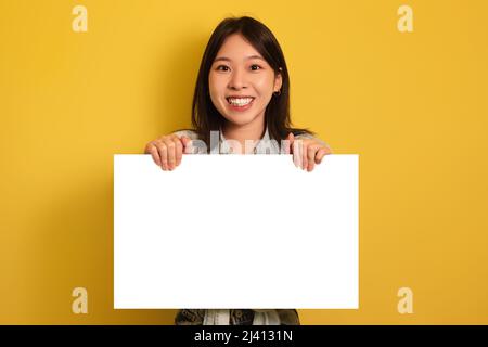 Glückliche junge asiatische Frau mit leerem Papierbanner, lächelnd auf Kamera auf gelbem Studiohintergrund, Mockup Stockfoto