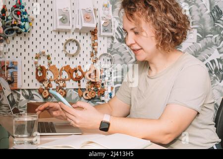 Lächelnde, konzentrierte Geschäftsfrau, die Smartphone spricht, am Laptop arbeitet, Ordnung nimmt. Babyzubehör, Silikon-Öko-Rasseln, Beruhigungssauger, Stockfoto