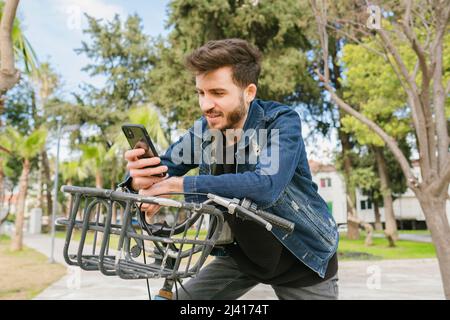 Junger Mann, der auf einem Leihfahrrad im Stadtpark telefonieren möchte. Er wartet auf seinen Freund. Nachrichten, Scrollen oder Ansehen von Videos. Videoanruf von seinem gi Stockfoto