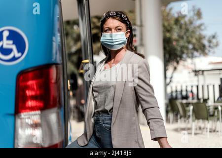 Eine junge Frau mit einer schützenden maedizinischen Maske, die mit einem Gepäck in den Bus einfährt Stockfoto