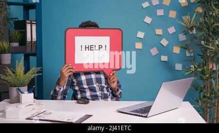 Ein männlicher Mitarbeiter hält eine Sprechblase mit einer Textnachricht, um Hilfe zu bitten, während er an einem Laptop arbeitet, um eine Geschäftsstrategie zu planen. Projektmanager zeigt Karteikarte mit Schrift auf Cartoon-Mockup. Stockfoto