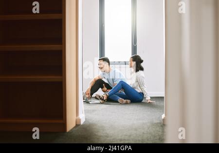 Wir haben in diesem Haus so viel zu tun. In voller Länge ein junges Paar, das im neuen Haus zusammen auf dem Boden sitzt und einen Laptop benutzt. Stockfoto