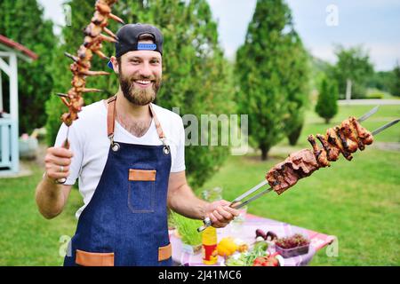 Der junge bärtige Mann in einer Mütze und einer Jeanschürze lächelt und hält einen Spieß mit köstlichem Grillfleisch, das vor dem Hintergrund auf dem Grill zubereitet wird Stockfoto