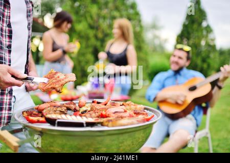 Die Hand des Mannes hält eine Grillzange mit einem saftig leckeren Fleischsteak vor dem Hintergrund eines Grillgrills mit Fleisch und Gemüse und einer Gruppe von Stockfoto