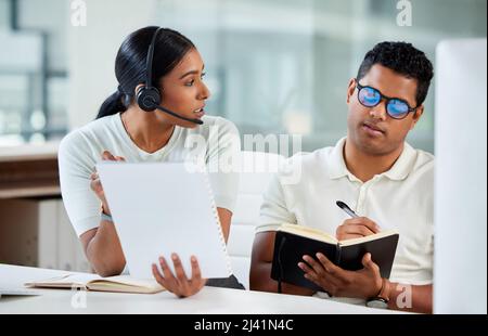 Machen Sie sich Notizen davon. Aufnahme einer attraktiven jungen Agentin, die mit ihrem Auszubildenden im Büro sitzt und erklärt, während er Notizen schreibt. Stockfoto