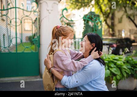 Glückliches Schulmädchen umarmt seine Großmutter wartet auf sie nach der Schule im Freien auf der Straße. Stockfoto