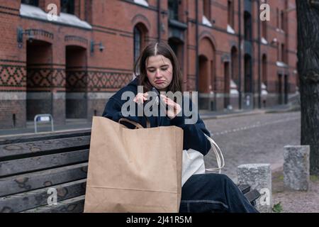 Eine junge Frau sitzt auf einer Bank mit einer leeren Brieftasche, Konzept des Mangels an Geld. Stockfoto