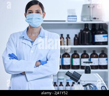 Ich werde Euch nicht im Stich lassen. Ein beschnittenes Porträt einer jungen Wissenschaftlerin, die eine Maske trägt, während sie mit gefalteten Armen in ihrem Labor steht. Stockfoto